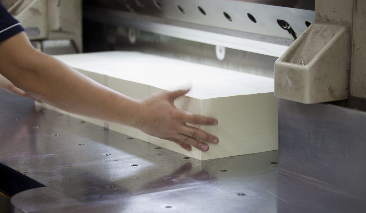 Books being trimmed with a heavy duty paper guillotine at a printing factory. Motion blur on the hands, selective focus.